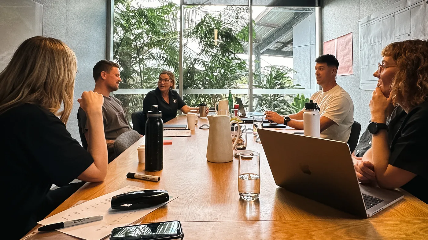 Seven people around a boardroom table in a session, palm trees visible through the window behind them