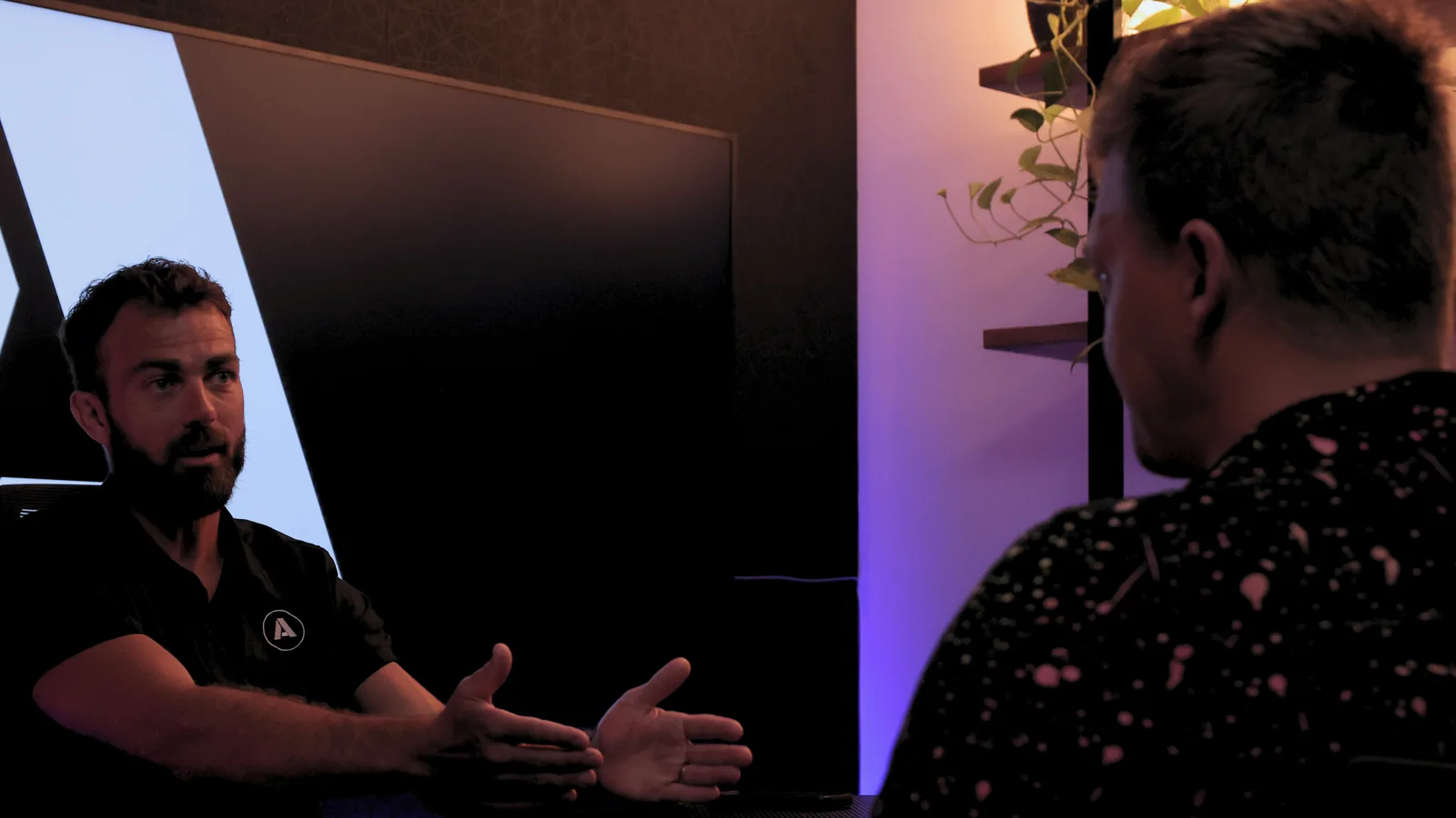 Greg Allan mid-explanation with his hands open, talking to a trade business owner across a desk in a session room