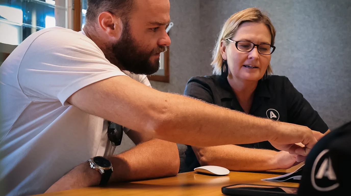 One owner reaching across the table to point at something on another owner's laptop screen, both of them looking at the same thing